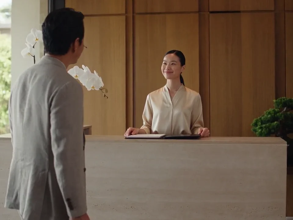 A hotel receptionist welcomes a guest at a light-filled reception desk.